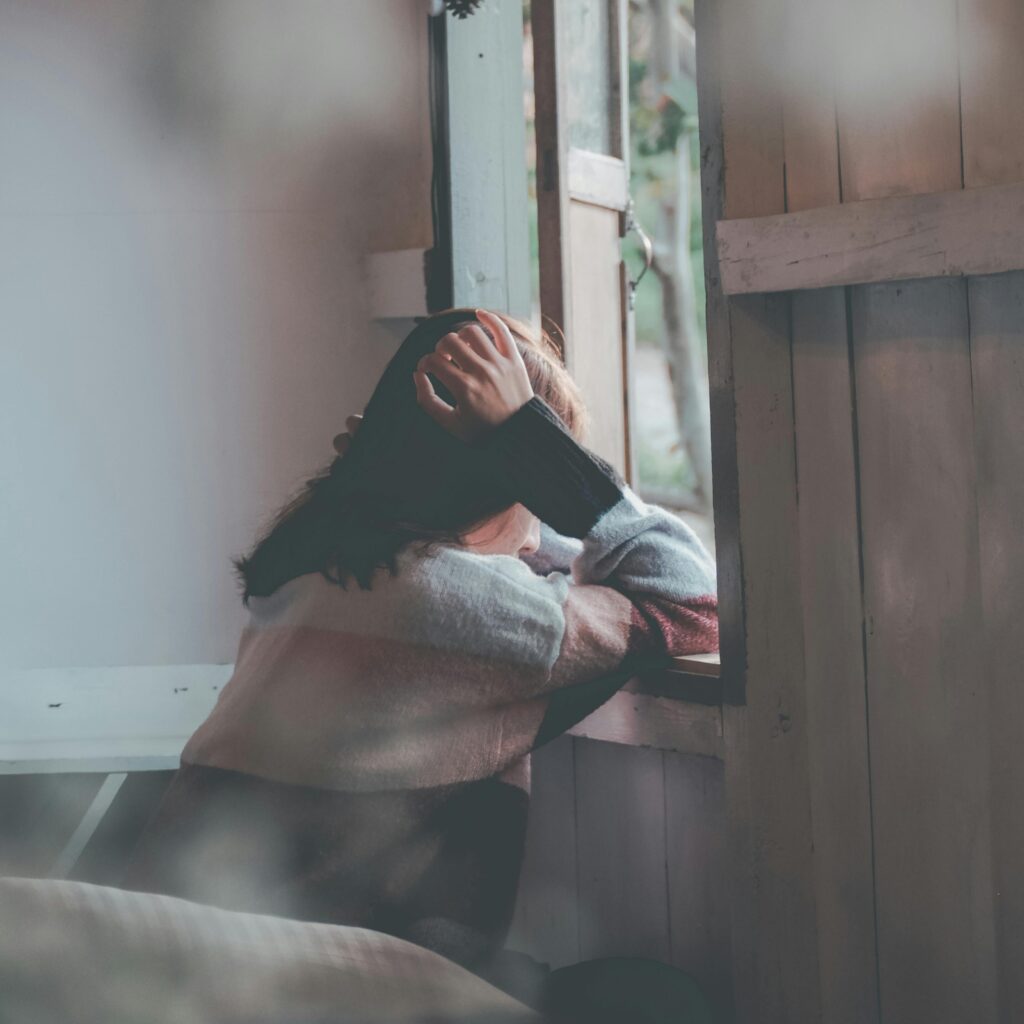 A woman sits indoors gazing thoughtfully out of a window, evoking emotions of loneliness and reflection.