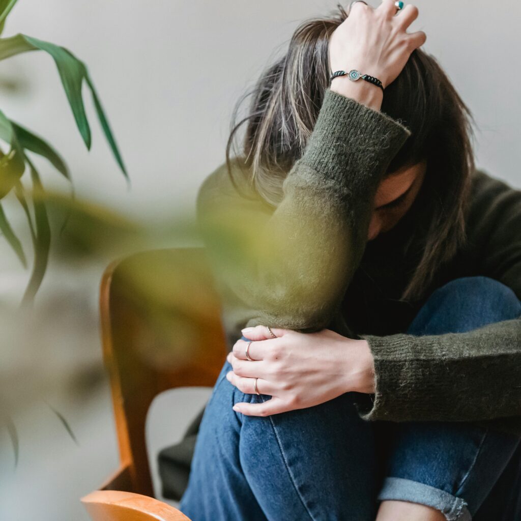 Anonymous young sorrowed female in casual outfit touching dark hair and embracing knees while sitting on chair at home