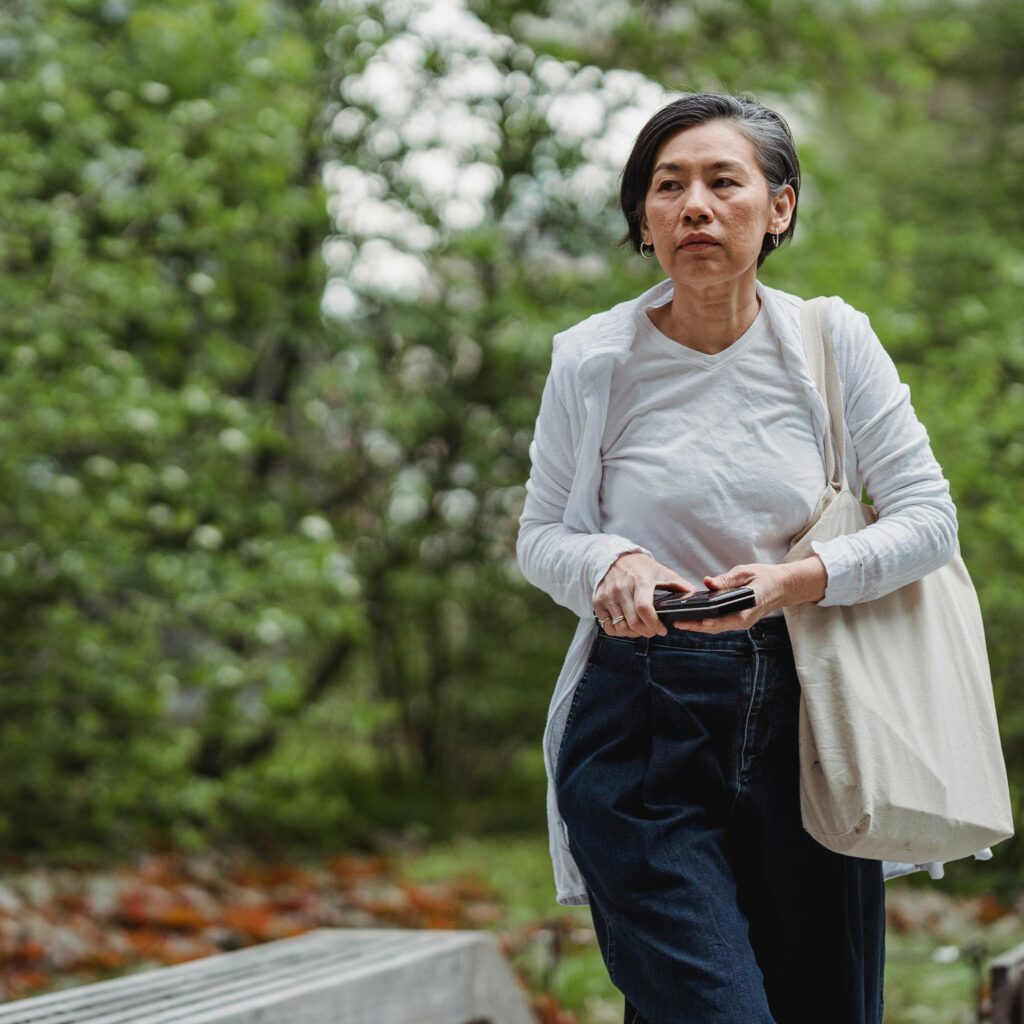 An adult woman walking outdoors with a phone and tote bag in hand. Casual and serene setting.