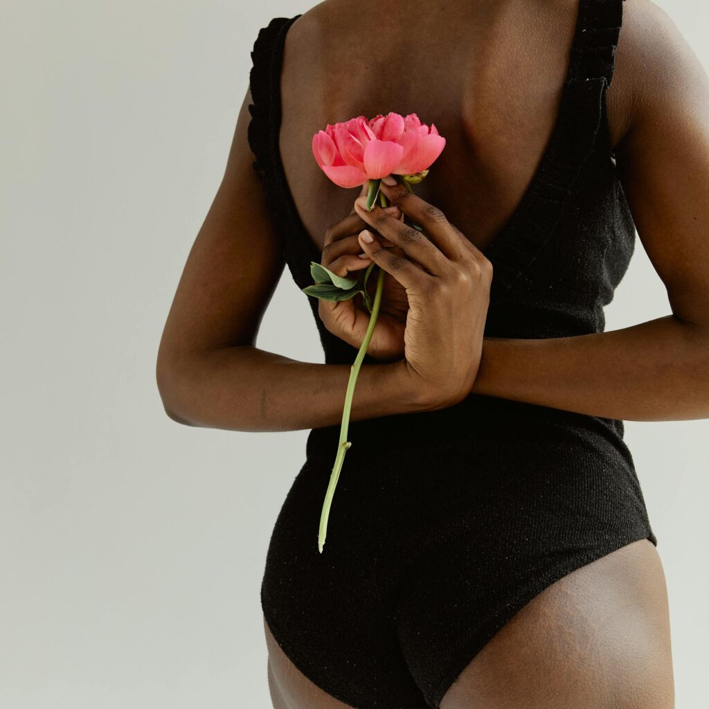 Aesthetic studio portrait of a woman in a black bodysuit holding a pink peony behind her back.